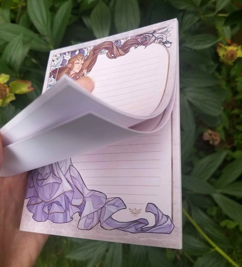 May include: A purple and white notepad with a decorative border featuring a woman with long brown hair and a purple dress. The notepad has lined pages and a gold Triforce symbol.
