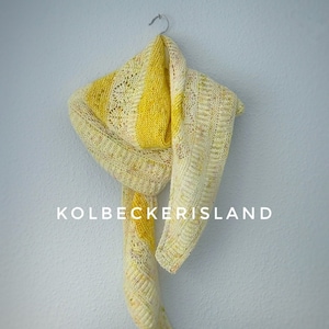 Può includere: Una sciarpa all'uncinetto a strisce gialle e bianche appesa a un gancio su una parete blu chiaro. Il testo "KOLBECKERISLAND" è stampato sotto la sciarpa.