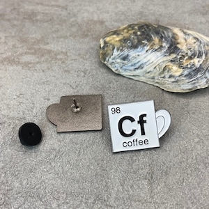 Könnte beinhalten: Eine weiße Emaille-Anstecknadel in Form einer Kaffeetasse mit dem chemischen Symbol "Cf" und dem Wort "coffee" darunter. Die Anstecknadel hat eine schwarze Rückseite und eine silberne Nadel.