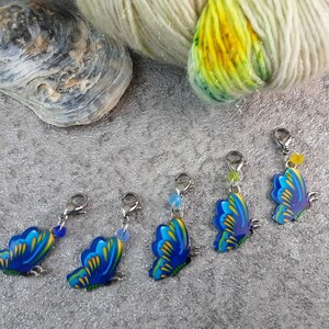 Könnte beinhalten: Fünf blaue und gelbe Schmetterlings-Maschenmarkierer mit silbernen Ringen und Perlen. Die Markierer befinden sich auf einer grauen, strukturierten Oberfläche.