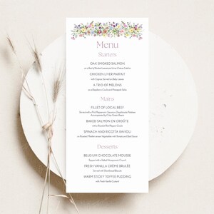 Może przedstawiać: Prostokątna karta menu z kwiatowym wzorem u góry, zawierająca listę dań. Menu zawiera przystawki, dania główne i desery. Karta jest biała i spoczywa na białej, okrągłej powierzchni.