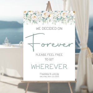 Op de afbeelding: Een wit huwelijksbord met groen blad en witte bloemen. Het bord staat "We decided on Forever. Please feel free to sit wherever. Freddie & Lottie. Saturday 26th July 2025."
