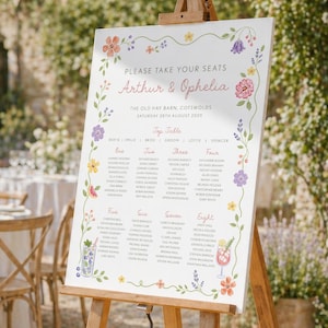Peut inclure: Un plan de table de mariage blanc sur un chevalet, orné d'illustrations florales roses, violettes, jaunes et oranges. Le plan affiche les noms et les affectations de table, avec le texte "Arthur & Ophelia" en haut. Le fond est un jardin.