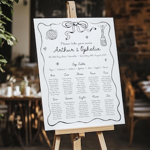 May include: A white wedding seating plan displayed on a wooden easel. The chart has black handwritten text, including the names "Arthur & Ophelia" and table assignments. Decorative elements include a disco ball, champagne bottle, and bow.