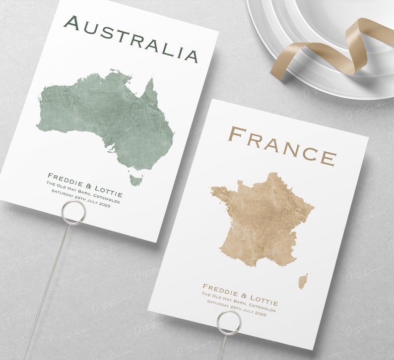 May include: Two place cards with a map of Australia and France. The cards are white with brown text and a green and brown map. The text reads "Freddie & Lottie" and "The Old Hay Barn, Cotswolds" and "Saturday 26th July 2025".