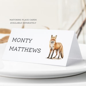 May include: A white place card with the name "MONTY MATTHEWS" printed in black text. A watercolor illustration of a red fox is on the right side of the card.
