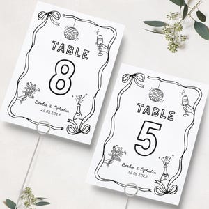 Könnte beinhalten: Zwei schwarz-weiße Tischkarten mit den Zahlen 8 und 5. Jede Karte hat einen dekorativen Rahmen, eine Discokugel, Champagner und den Text „TABLE“. Die Namen „Bertie & Ophelia“ und ein Datum sind ebenfalls enthalten.