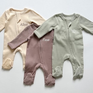 May include: Three ribbed baby footie pajamas in beige, mauve, and sage green. Each has a zipper closure and the name "Albie", "Ezra", or "Smith" embroidered on the chest. The footies have textured soles.