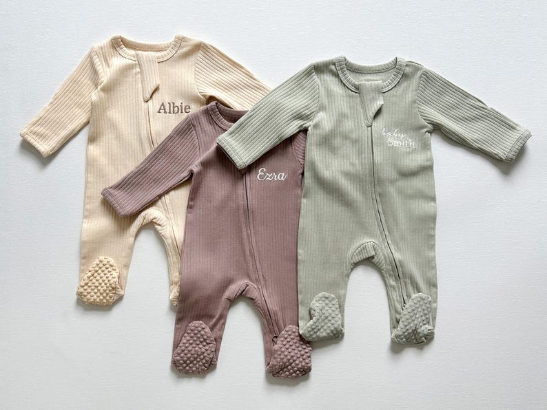 May include: Three ribbed baby footie pajamas in beige, mauve, and sage green. Each has a zipper closure and the name "Albie", "Ezra", or "Smith" embroidered on the chest. The footies have textured soles.