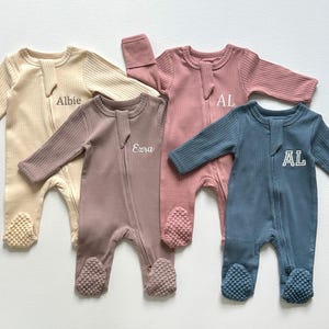 May include: Four ribbed cotton baby sleepers in beige, mauve, pink, and blue. Each sleeper has a zipper closure, footed bottoms, and embroidered text or initials. The sleepers are designed for infants and toddlers.