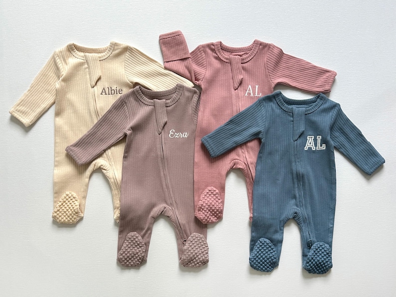 May include: Four ribbed cotton baby sleepers in beige, mauve, pink, and blue. Each sleeper has a zipper closure, footed bottoms, and embroidered text or initials. The sleepers are designed for infants and toddlers.