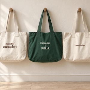 May include: Three canvas tote bags hanging on a wall. The left bag is off-white with the text "custom embroidery" in brown. The centre bag is dark green with the text "Expensive & Difficult" in white. The right bag is off-white with "manifesting." in brown.
