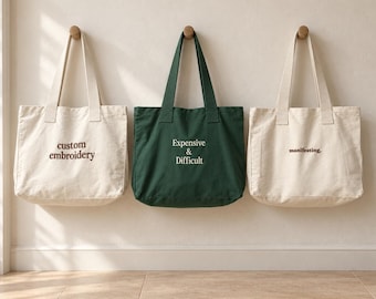 May include: Three canvas tote bags hanging on a wall. The left bag is off-white with the text custom embroidery in brown. The centre bag is dark green with the text Expensive & Difficult in white. The right bag is off-white with the text manifesting. in brown.