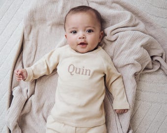 Könnte beinhalten: Ein Baby in einem hellbeigen Langarmshirt und Hosen-Set. Auf der Vorderseite des Shirts ist der Name "Quin" aufgestickt. Das Baby liegt auf einer beigen Decke.