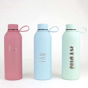 May include: Three matte finish water bottles in dusty rose, light blue, and mint green. Each bottle features a matching screw-top lid with a flexible silicone carrying loop and a silver-toned engraved name on the front: Zaria, AVI, and AYDIN.