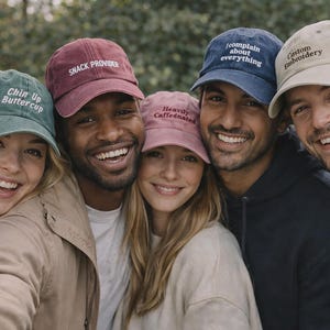 May include: A group of smiling people wearing various colored baseball caps. The caps are in shades of green, burgundy, pink, navy blue, and tan. Each cap has white embroidered text, including phrases like "Chin Up Buttercup" and "Custom Embroidery."