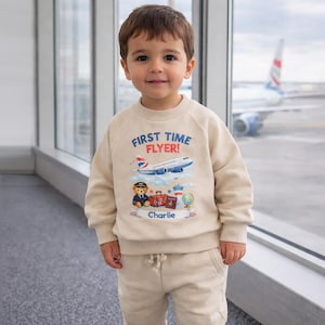 Könnte beinhalten: Ein Kind trägt einen beigefarbenen Pullover und eine passende Hose. Der Pullover zeigt den Text "FIRST TIME FLYER!" über einer Flugzeuggrafik, einem Teddybären in Pilotenuniform und Illustrationen zum Thema Reisen. Der Name des Kindes, "Charlie", ist ebenfalls aufgedruckt.