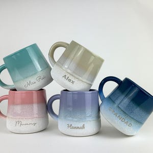 Könnte beinhalten: Fünf Keramikbecher in einem Stapel, jeder in einer anderen Farbe: Türkis, Rosa, Beige, Lila und Blau. Die Becher haben einen weißen Sockel und einen Farbverlauf. Auf den Bechern stehen die Namen "Mrs Rai", "Alex", "Mummy", "Hannah" und "Grandad".