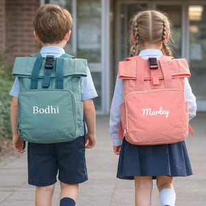 Può includere: Due bambini con zaini. Uno è verde acqua con il nome "Bodhi" in corsivo bianco, l'altro è corallo con "Marley". Entrambi hanno un design a rotolo. I bambini indossano uniformi scolastiche.