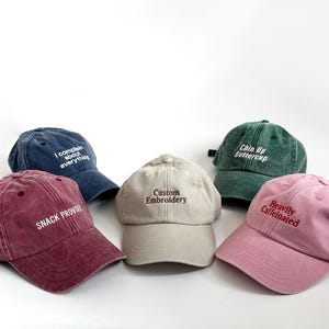 May include: Five baseball caps in various colors: burgundy, navy, beige, green, and pink. Each cap features embroidered text: "SNACK PROVIDED", "I complain about everything", "Custom Embroidery", "Chin Up Buttercup", and "Heavily Caffeinated".