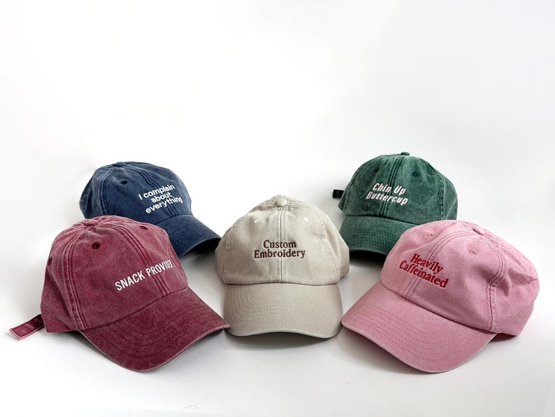 May include: Five baseball caps in various colors: burgundy, navy, beige, green, and pink. Each cap features embroidered text: "SNACK PROVIDED", "I complain about everything", "Custom Embroidery", "Chin Up Buttercup", and "Heavily Caffeinated".