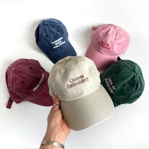 May include: A selection of baseball caps in shades of navy, pink, burgundy, green, and beige. Each cap has embroidered text, including phrases like "I complain about everything," "Heavily caffeinated," "Snack Provider," "Custom Embroidery," and "Chin Up Buttercup."