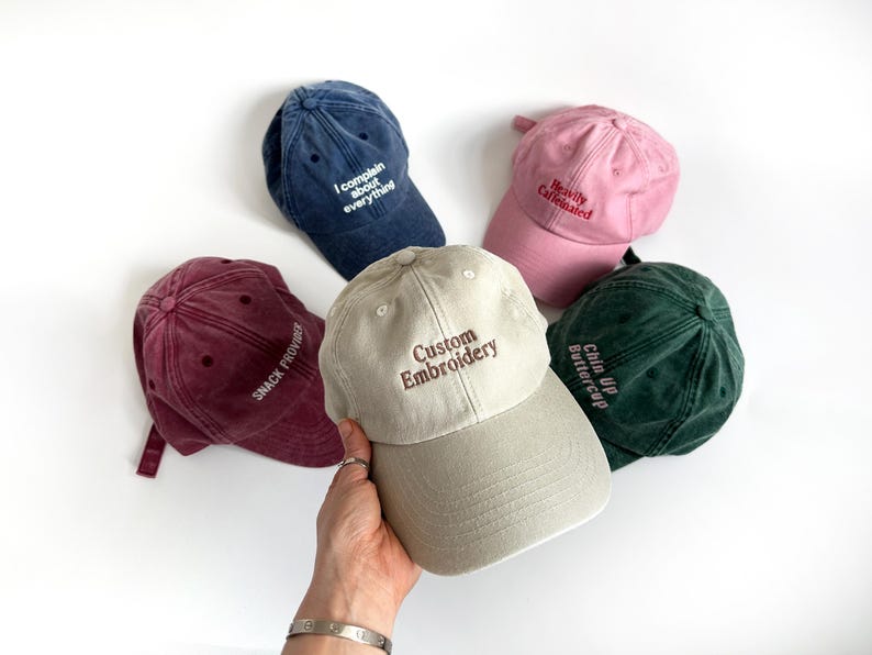 May include: A collection of five baseball caps in various colors: navy blue, pink, burgundy, green, and tan. Each cap features embroidered text, including "I complain about everything," "Heavily Caffeinated," "Snack Provider," "Custom Embroidery," and "Chin Up Buttercup."