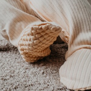 May include: Close-up of cream-colored baby footed pajamas. The ribbed fabric is visible on the legs, with textured, padded soles. The pajamas are designed for comfort and warmth. The soft, neutral color makes them suitable for any baby.