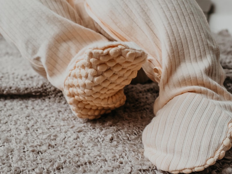 May include: Close-up of cream-colored baby footed pajamas. The ribbed fabric is visible on the legs, with textured, padded soles. The pajamas are designed for comfort and warmth. The soft, neutral color makes them suitable for any baby.