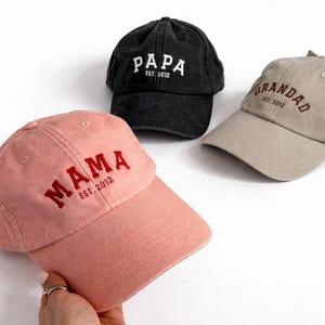 May include: Three baseball caps in pink, black, and beige. The pink cap features red embroidery reading MAMA EST. 2012. The black cap has white embroidery reading PAPA EST. 2012. The beige cap has brown embroidery reading GRANDAD EST. 2012.