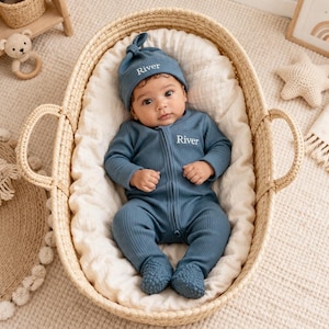 May include: A baby wearing a blue ribbed knit jumpsuit and matching knotted hat, both embroidered with the name "River". The baby is lying in a woven basket lined with a white blanket. The image has a soft, neutral color palette.