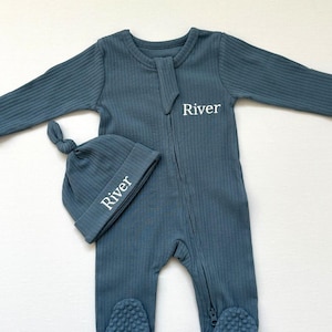 May include: A teal-colored baby sleeper and matching knotted hat set. The sleeper has a ribbed texture, a zipper closure, and foot coverings. The hat and sleeper are embroidered with the name "River" in white. This set is designed for newborns.