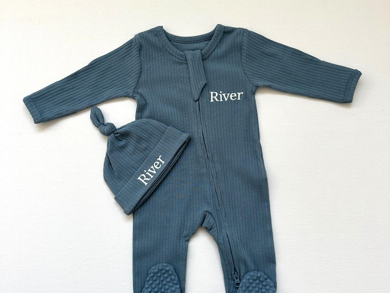May include: A teal-colored baby sleeper and matching knotted hat set. The sleeper has a ribbed texture, a zipper closure, and foot coverings. The hat and sleeper are embroidered with the name "River" in white. This set is designed for newborns.