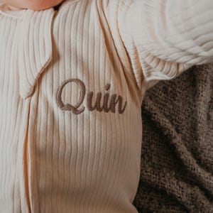 May include: Cream-colored, ribbed baby onesie with the name "Quin" embroidered in a soft brown thread. The onesie has a zipper closure and long sleeves. The garment is lying on a textured, brown blanket.