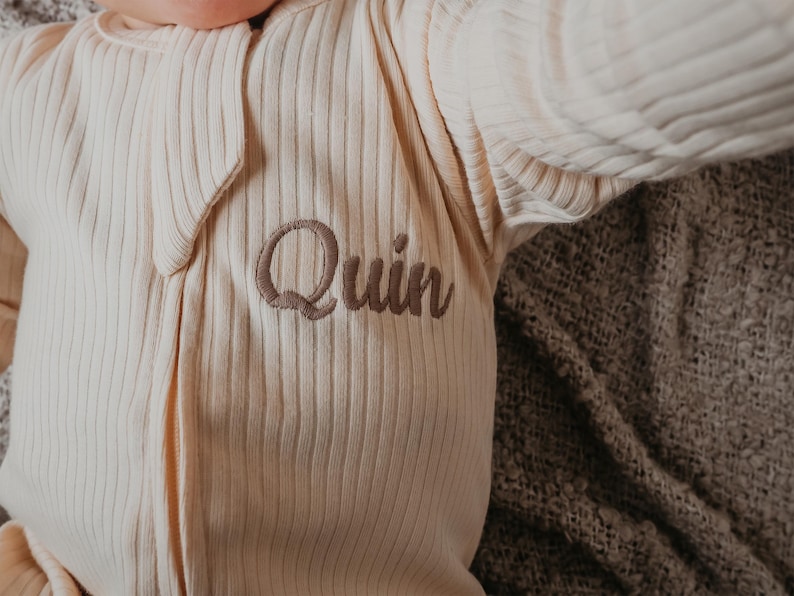 May include: Cream-colored, ribbed baby onesie with the name "Quin" embroidered in a soft brown thread. The onesie has a zipper closure and long sleeves. The garment is lying on a textured, brown blanket.