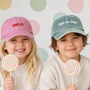 May include: Two children wearing baseball caps and holding lollipops. One cap is pink with "AMELIA" embroidered in red. The other is teal with "High on Sugar" embroidered in white. The lollipops are swirled with pastel colours.