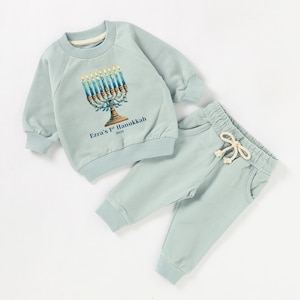 May include: A light blue baby sweatshirt with a menorah graphic and the text "Ezra's 1st Hanukkah 2023". The sweatshirt is paired with matching light blue sweatpants.