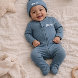 May include: A baby wearing a blue ribbed knit footed onesie and matching knotted hat, both embroidered with the name "River". The baby is smiling and lying on a cream-colored blanket. A star-shaped pillow and a wooden toy are in the background.