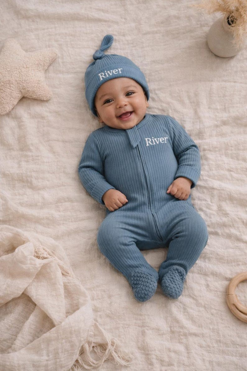 May include: A baby wearing a blue ribbed knit footed onesie and matching knotted hat, both embroidered with the name "River". The baby is smiling and lying on a cream-colored blanket. A star-shaped pillow and a wooden toy are in the background.