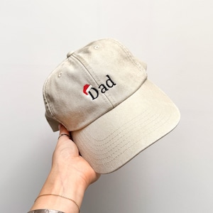 May include: A beige baseball cap featuring embroidered black text that reads Dad, with a small red and white Santa hat icon positioned directly above the letter D. The cap has a structured crown, a curved brim, and visible stitching details.