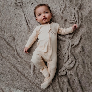 May include: A baby wearing a light yellow, long-sleeved, zippered sleeper with the name "Quinn" embroidered on the front. The baby is lying on a brown, fuzzy blanket.