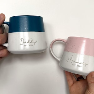 Puede incluir: Dos tazas de cerámica, una verde azulado y blanca, la otra rosa y blanca. La taza verde azulado dice "Daddy EST. 2026" y la taza rosa dice "Mummy EST. 2026". Ambas tazas tienen un acabado moteado.