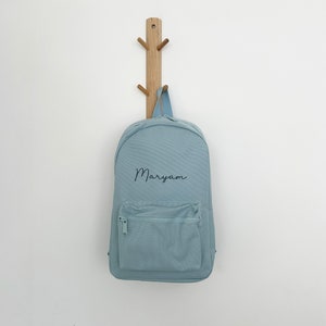 May include: A light blue backpack with a front zippered pocket and the name "Maryam" in script. The backpack is hanging on a wooden coat rack against a white wall.
