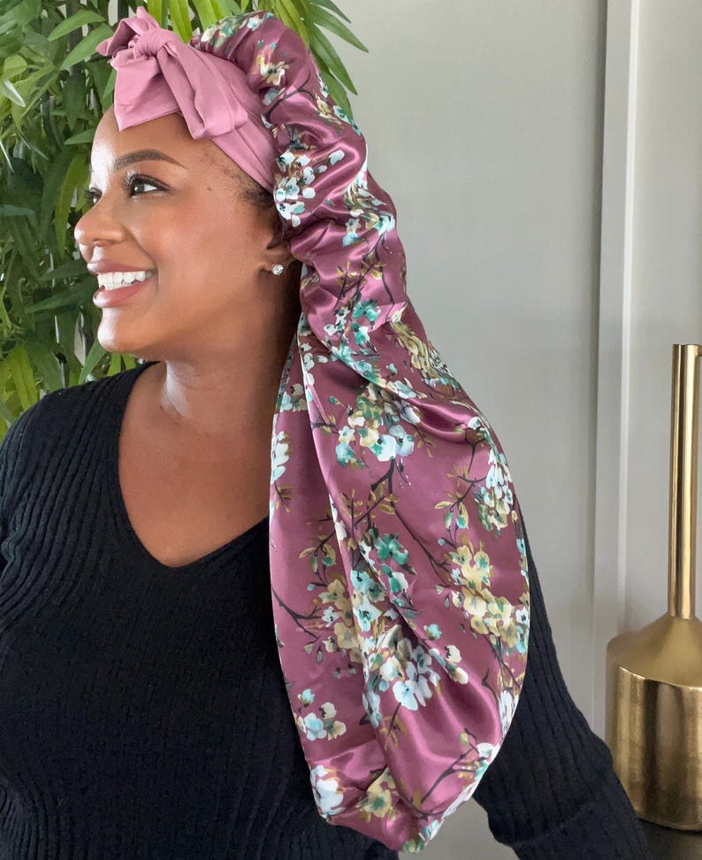 May include: A pink satin sleep cap with a floral print. The cap has a bow on top and is designed to protect hair while sleeping.