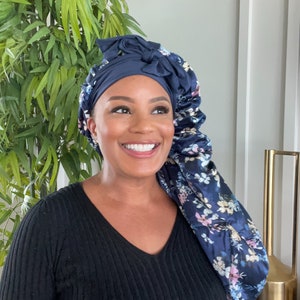 May include: A woman wearing a navy blue satin sleep cap with a floral print. The cap has a bow on top and is tied at the back of the head. She is wearing a black sweater.