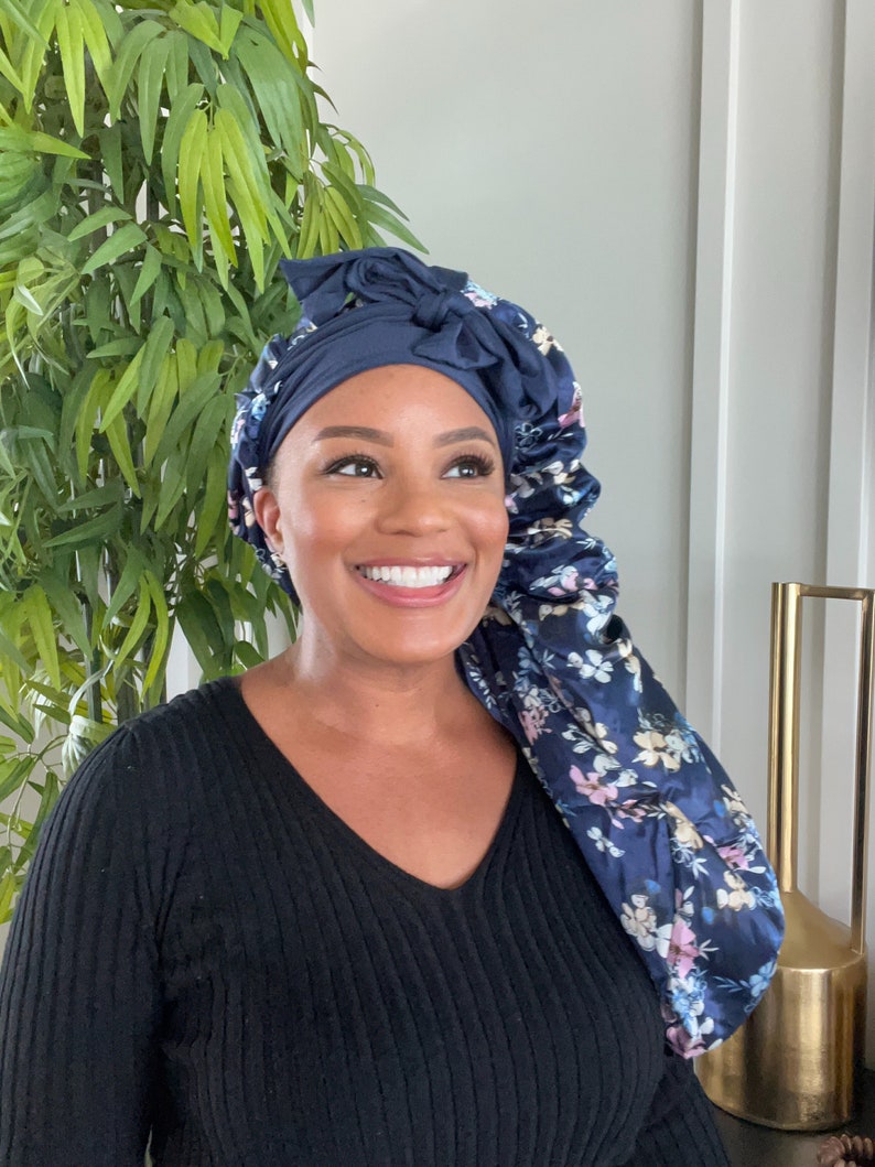 May include: A woman wearing a navy blue satin sleep cap with a floral print. The cap has a bow on top and is tied at the back of the head. She is wearing a black sweater.