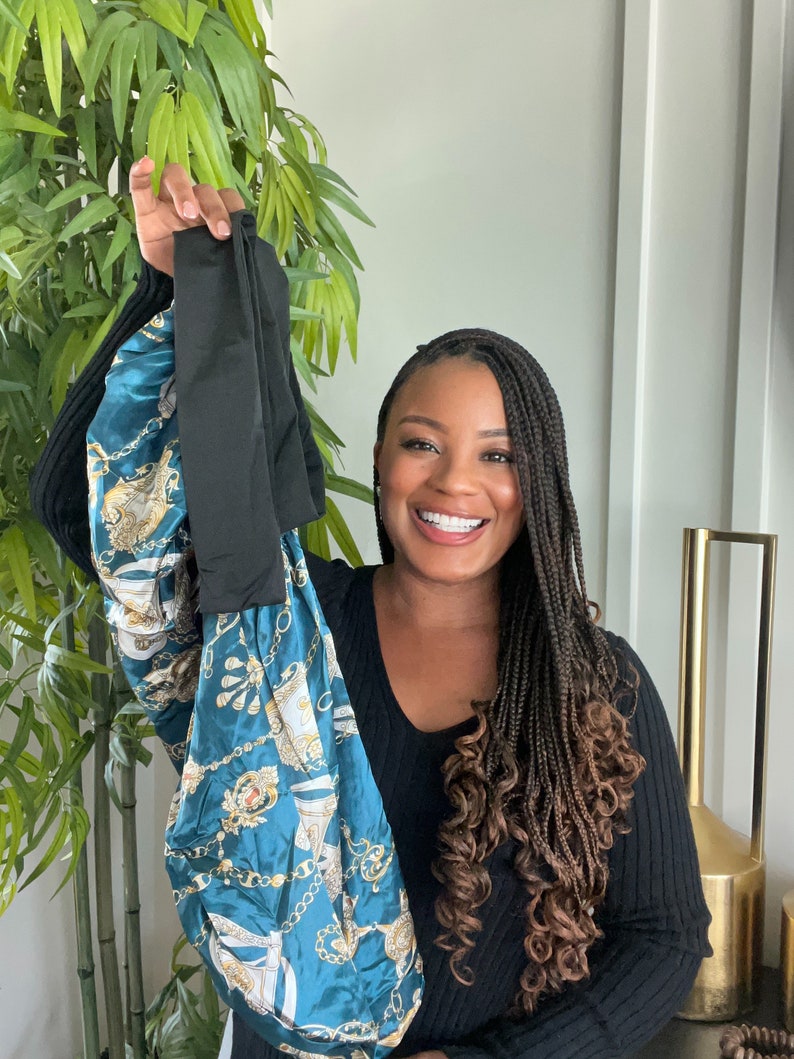 May include: A woman with long braids is holding up a teal blue and gold satin bag with a black fabric handle. The bag has a pattern of chains and ornate designs.