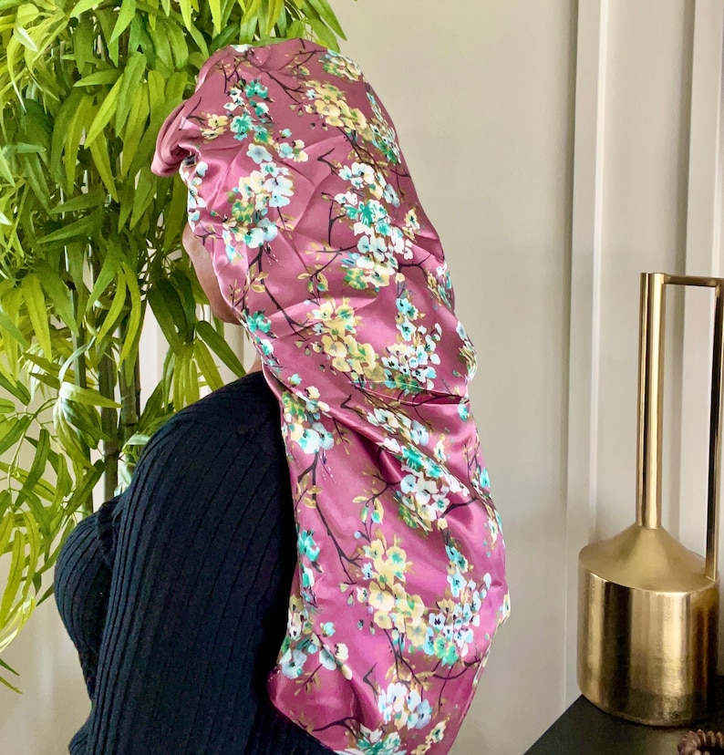 May include: A pink satin sleep cap with a floral print featuring white, yellow, and green flowers. The cap is designed to protect hair while sleeping.