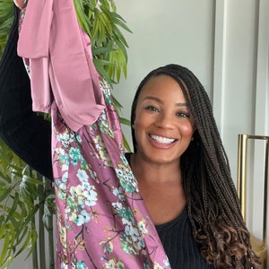 May include: A woman with long black braids is holding up a pink and floral patterned dress. The dress has a floral print with pink, green, and white flowers.