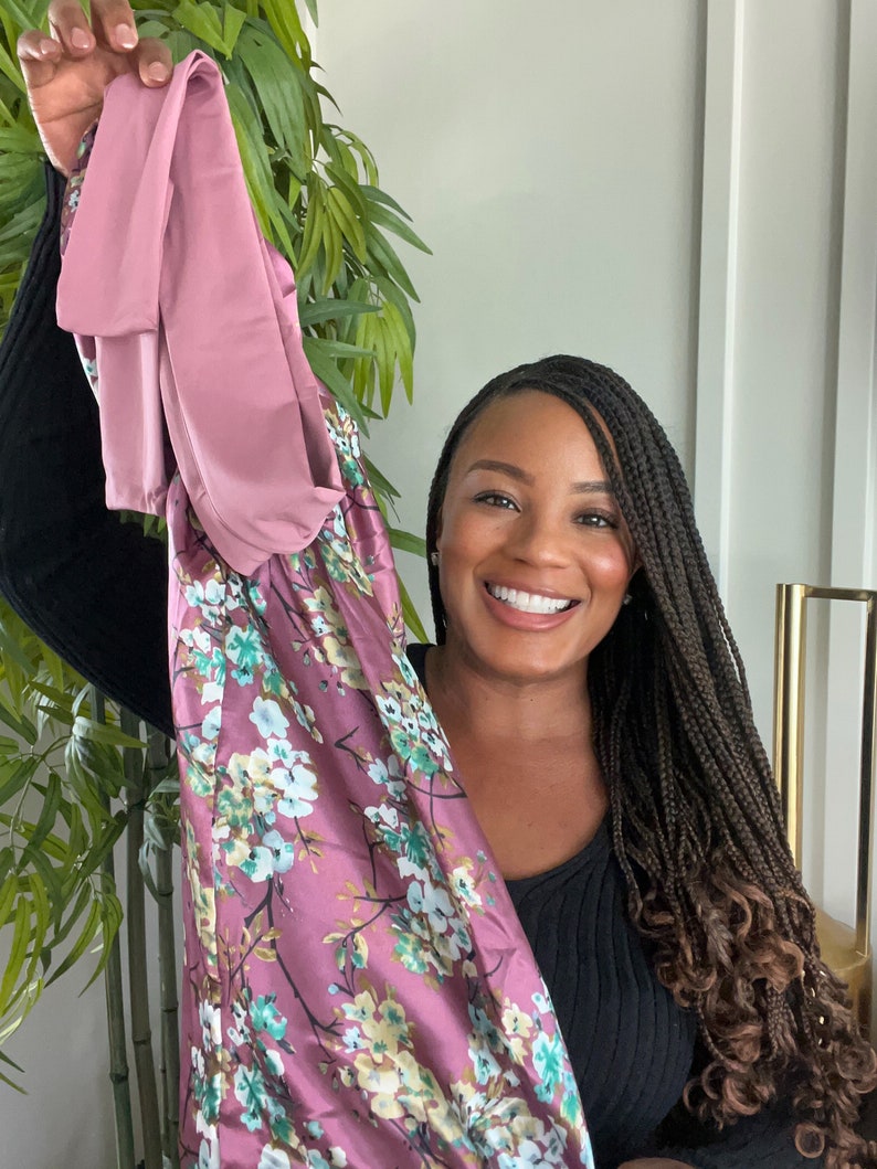 May include: A woman with long black braids is holding up a pink and floral patterned dress. The dress has a floral print with pink, green, and white flowers.